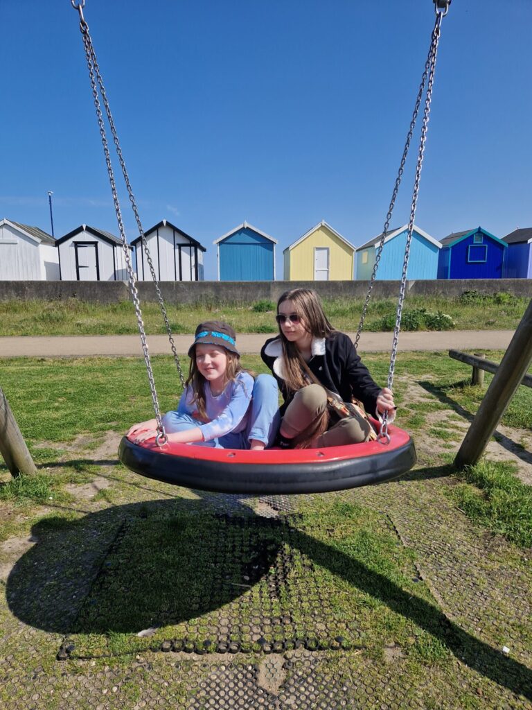 Tigerboy and Smallest on saucer swing with beach huts in background and blue sky