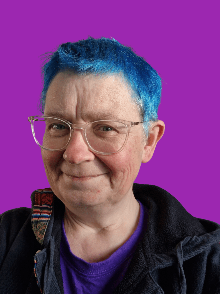 Jax, a non binary individual with short blue hair and clear framed plastic glasses smiles at camera. They are wearing a deep blue hooded jumper with woven fabric around the neckline.
