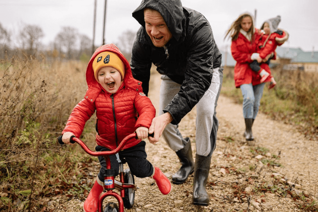 Small child on bike smiling widely, man leaning down and pushing the bike along